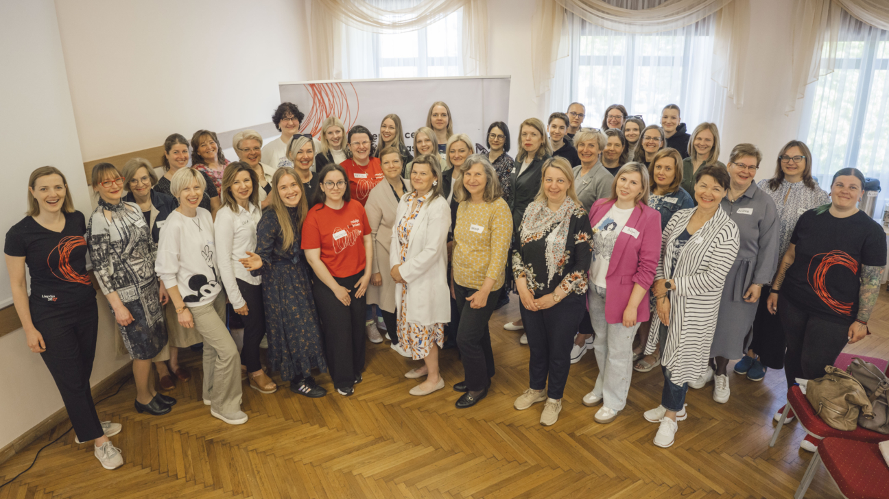 Liela personu grupa pozē kopīgai fotogrāfijai iekštelpās uz koka grīdas fona, daļa no personām valkā Liepāja 2027 krekliņus vai sarkanus T-kreklus ar grafisku dizainu. Aizmugurē redzams baneris ar Liepāja 2027 vizuālajiem elementiem un gaiši aizkari pie logiem.