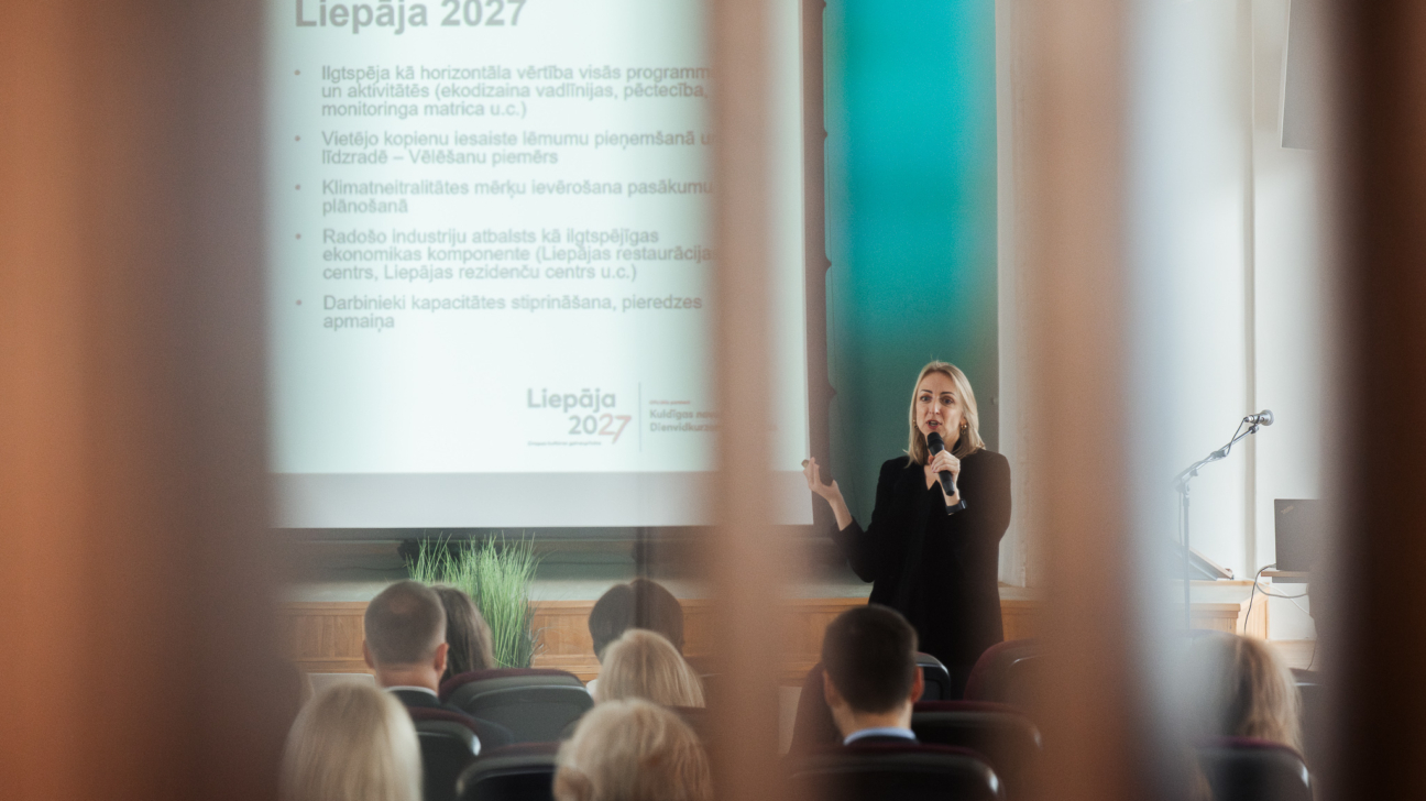 Priekšplānā semināra lektors, uz ekrāna tiek rādīta prezentācija