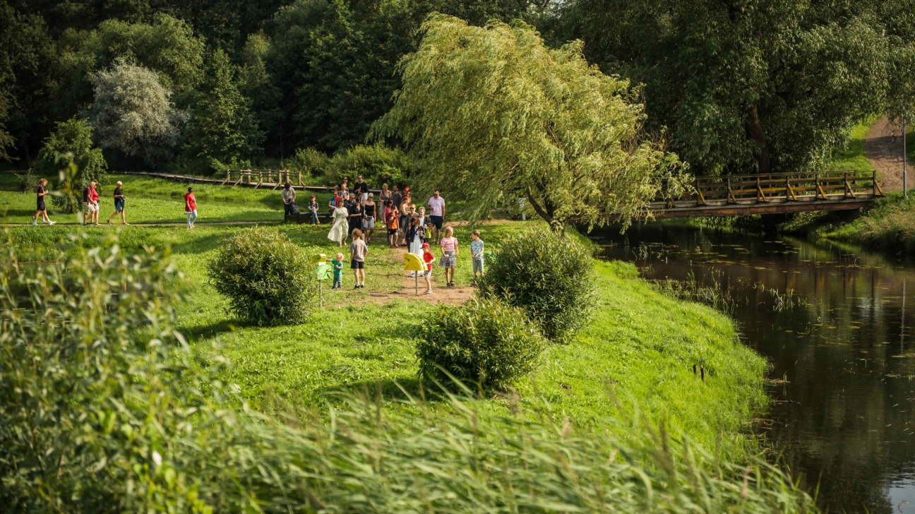 Cilvēku grupa, tostarp bērni, pulcējusies uz zāļaina krasta pie upes parkā, daži staigā pa taku. Koka tilts šķērso upi, ko ieskauj blīvi koki.