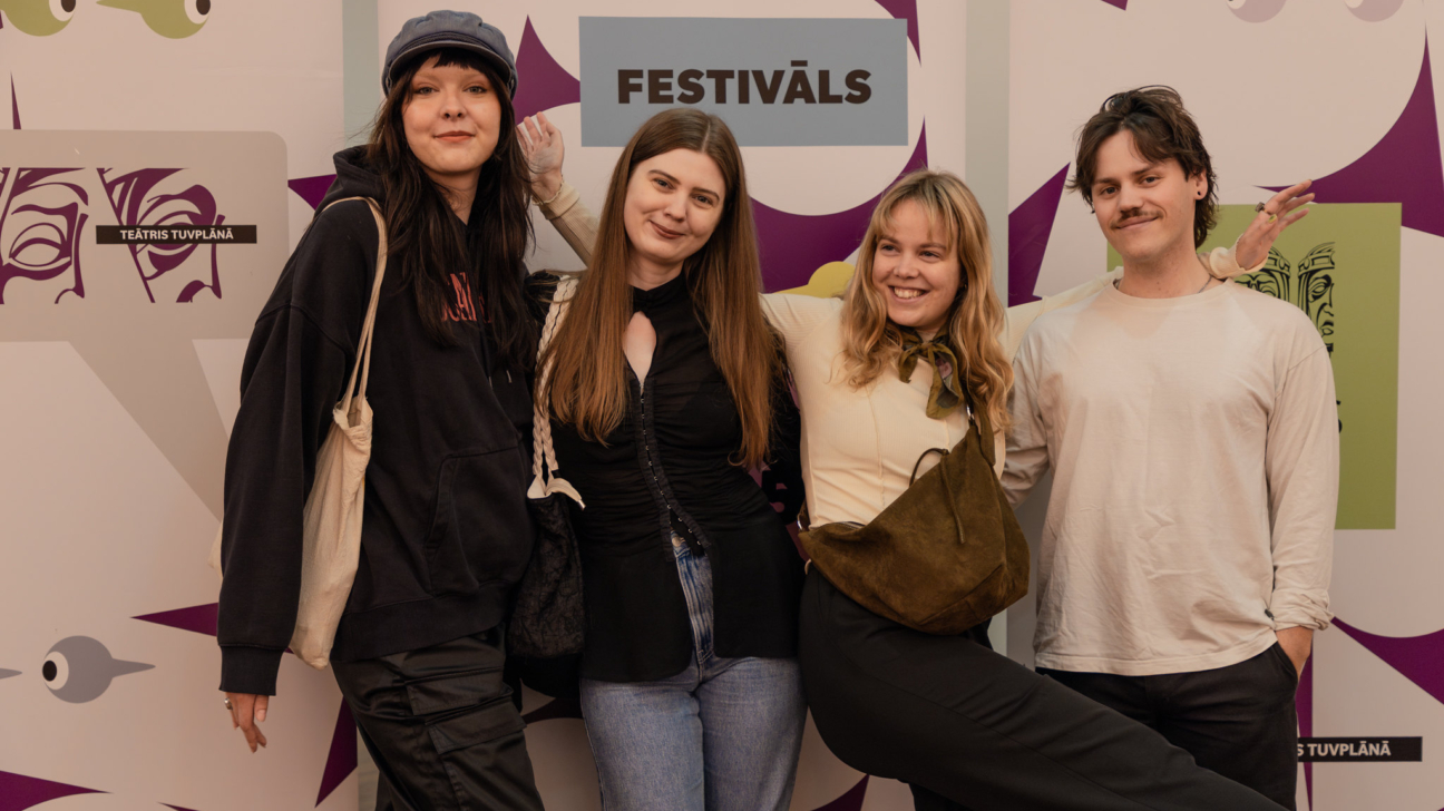 Četri jauni cilvēki smaidīgi pozē kopīgā fotogrāfijā uz festivāla 'Teātris tuvplānā' fona. No kreisās – sieviete melnā džemperī ar kapuci un auduma somu uz pleca, blakus viņai stāv sieviete melnā blūzē ar džinsiem, trešā sieviete gaišā blūzē ar lakatiņu ap kaklu un lielu somu pāri plecam smaidot izpleš rokas, bet ceturtais ir vīrietis gaišā kreklā ar ūsām, kurš smaida kamerai. Fonā redzams uzraksts 'Festivāls' un stilizētas teātra masku ilustrācijas violetā un pelēkā tonī."

Vai vēlies, lai izveidoju arī īso alt tekstu tīmekļa pieejamī