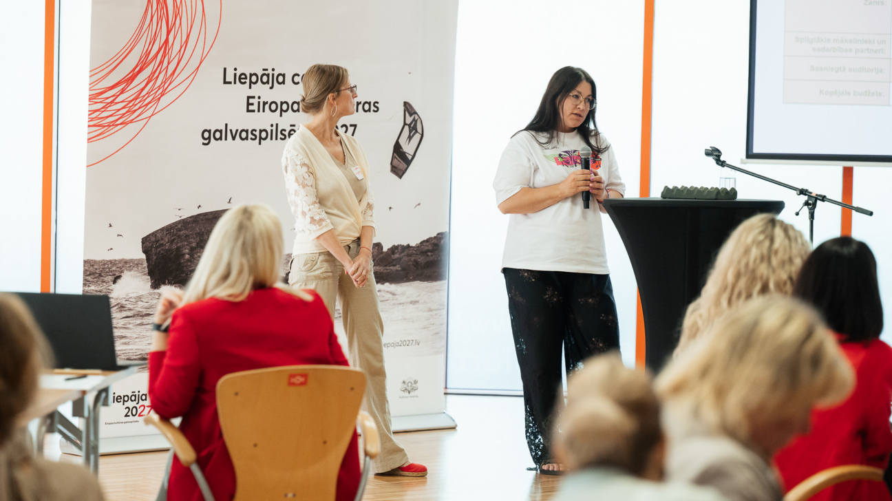 Divas sievietes prezentē auditorijai pasākumā, kas saistīts ar Eiropas kultūras galvaspilsētu 2027. Viena sieviete, ģērbusies gaišā kostīmā ar mežģīņu piedurknēm, stāv pie plakāta ar uzrakstu 'Liepāja ceļā uz Eiropas kultūras galvaspilsētu 2027'. Otrā sieviete, melniem matiem un brillēm, ģērbusies baltā T-kreklā un melnās biksēs, runā mikrofonā pie melna stenda ar mikrofonu statīvā. Auditorijā redzamas vairākas sēdošas personas