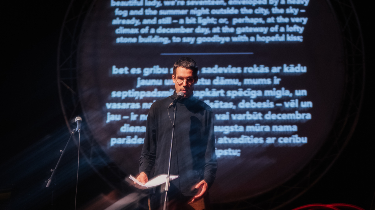 Jauns vīrietis ar brillēm stāv uz skatuves pie mikrofona, turot rokās lapas ar tekstu. Fonā uz ekrāna projicēts literārs teksts angļu un latviešu valodā, radot vizuāli spēcīgu fona noskaņu. Skatuves apgaismojums veido māksliniecisku atmosfēru ar gaismas efektiem, kas piešķir notikumam emocionālu un poētisku noskaņu. Attēls ataino dzejas vai literāro lasījumu pasākumu