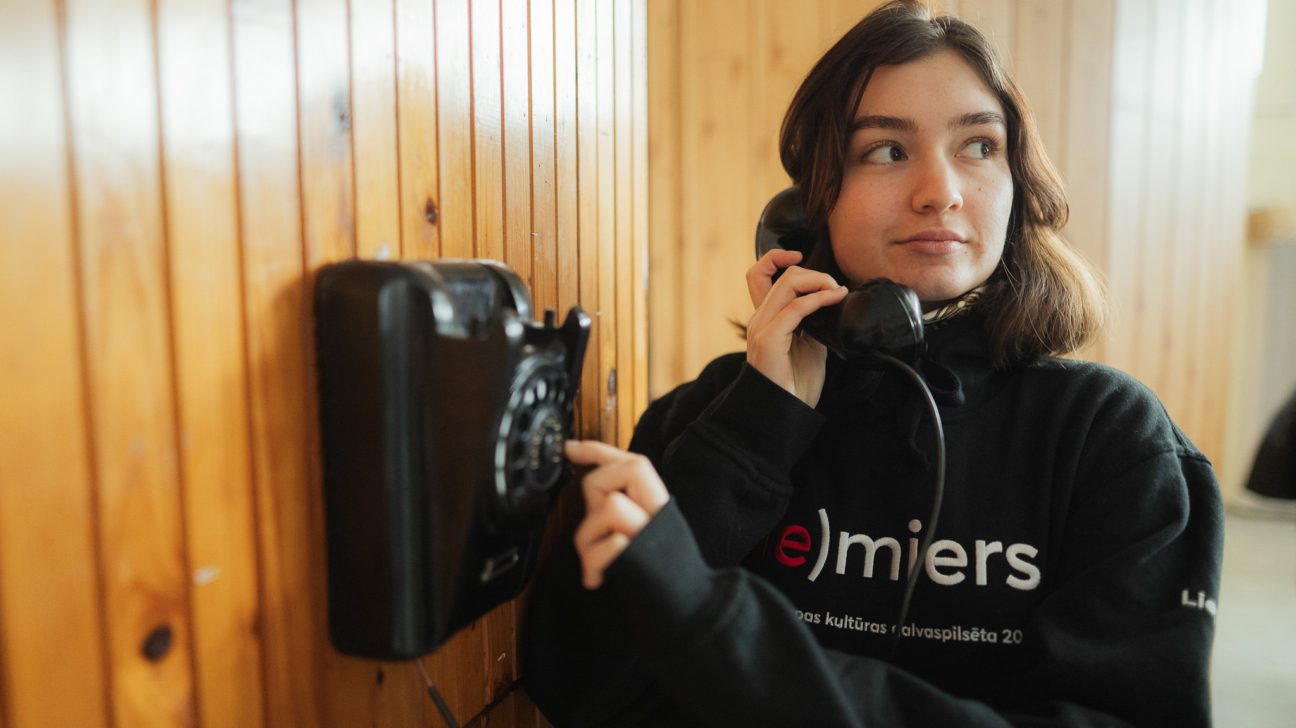 Jauna sieviete melnā džemperī ar uzrakstu “(e)miers” runā pa vecmodīgu melnu diska telefonu pie koka sienas