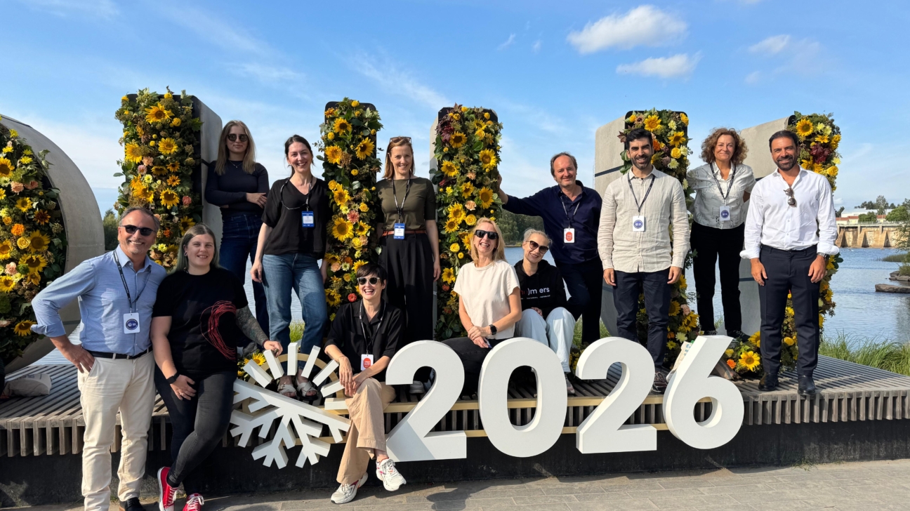 Cilvēku grupa pozē pie liela uzraksta '2026', kas novietots ārpus telpām pie ūdens. Skaitlis papildināts ar sniegpārslas simbolu, bet aiz tā redzamas dekorācijas no spilgti dzelteniem saulespuķu un citu ziedu kompozīcijām. Daļa cilvēku stāv, daļa sēž uz konstrukcijas malas, visi smaida un ir tērpušies neformālā, bet kārtīgā apģērbā ar pasākuma identifikācijas kartēm kaklā. Fonā redzama upe, tilts un zilas debesis ar mākoņiem