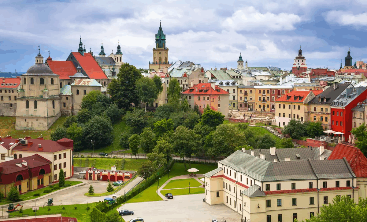 Krāsaina panorāmas ainava, kas ataino Ļubļinas (Polija) vecpilsētu ar tās daudzveidīgo arhitektūru. Attēlā redzamas krāsainas vēsturiskas ēkas ar sarkaniem, dzelteniem un pasteļtoņiem krāsotiem jumtiem, kā arī baznīcu torņi ar krustiem un zaļiem jumtiem, kas dominē pilsētas ainavā. Priekšplānā redzami koki, parki un pagalmi ar automašīnām un sporta laukumu, kas rada kontrastu starp zaļo vidi un pilsētas vēsturisko apbūvi. Fonā mākoņains, bet gaišs debesu fons, kas piešķir ainavai dzīvīgumu un dziļumu.