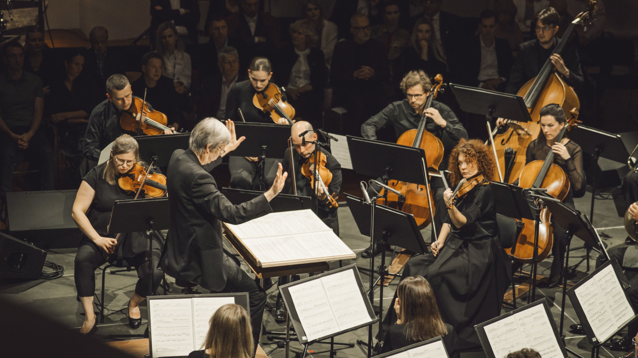 Orķestra koncerts ar diriģentu priekšplānā, kurš ar paceltām rokām vada mūziķus. Muzikanti spēlē vijoles, čellus un kontrabasu, tērpti melnā koncertapģērbā. Viņu priekšā atrodas notis uz statīviem, un fonā redzama uzmanīga skatītāju auditorija. Skatuves apgaismojums ir silts un koncentrēts uz orķestri, radot klasiskā koncerta cienīgu atmosfēru ar profesionālu noskaņu.