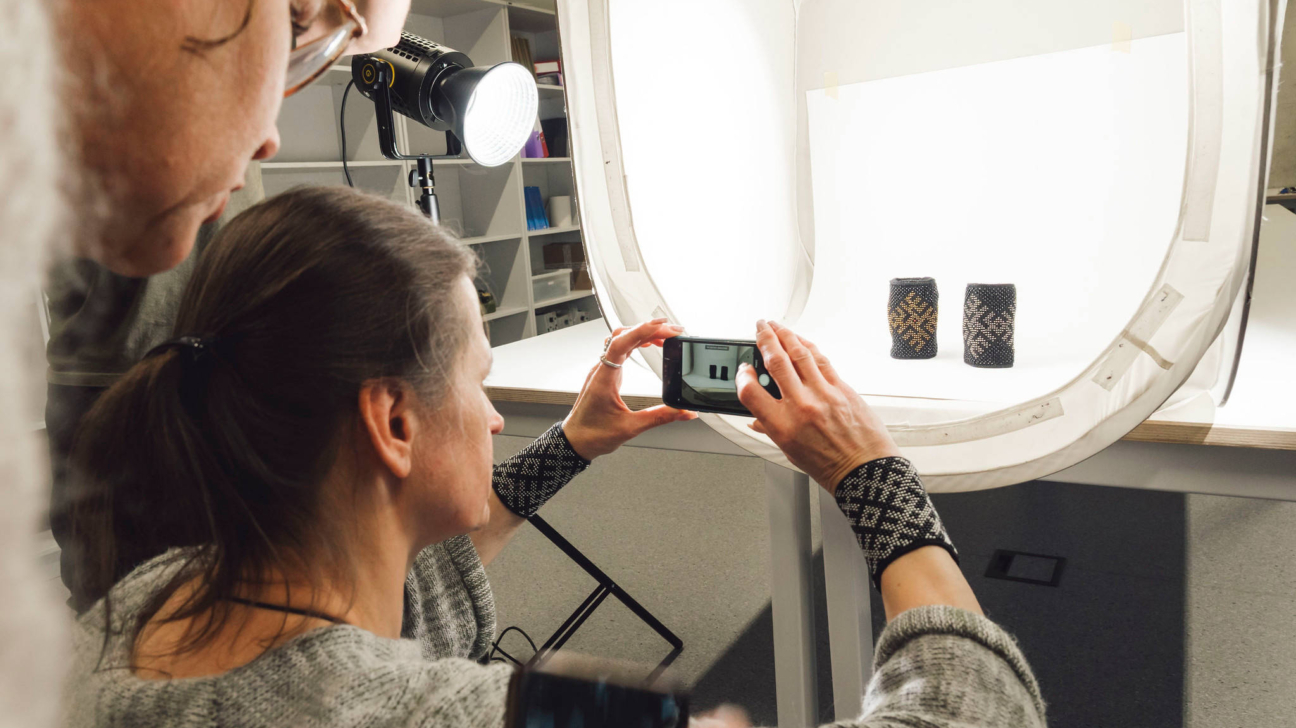 Attēlā redzami cilvēki, kas praktiski apgūst produktu fotografēšanas prasmes. Sieviete priekšplānā ar viedtālruni fotografē divus rokdarbus — cimdu pāri ar etnogrāfiskiem rakstiem —, kas novietoti baltā foto gaismas kastē ar spilgtu apgaismojumu no lampas. Fonā redzamas plauktu rindas ar materiāliem un aprīkojumu. Attēls ataino radošu un praktisku mācību procesu, kur dalībnieki iepazīst fotogrāfijas un digitālā satura veidošanas tehniku.