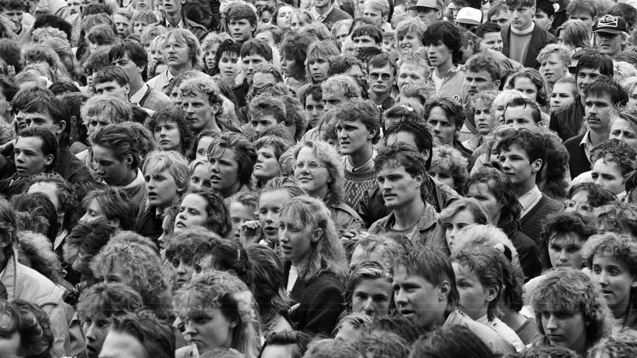 Vēsturiska melnbalta fotogrāfija, kurā redzams liels cilvēku pūlis brīvdabas pasākumā, visticamāk koncertā Liepājā 1980. gadu beigās. Attēlā redzami jaunieši un pieaugušie ar dažādiem to laika frizūru un apģērbu stiliem, kas uzmanīgi un emocionāli vēro notiekošo uz skatuves. Fonā redzamas cieši stāvošas cilvēku rindas, dažiem rokās ir lietussargi, radot sajūtu par blīvi piepildītu un nozīmīgu sabiedrisku notikumu padomju laika atmosfērā.