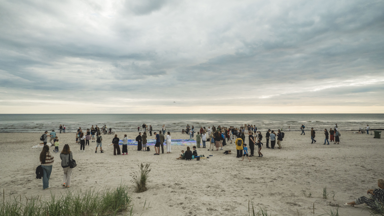 Cilvēku pulcēšanās Liepājas pludmalē pie Baltijas jūras mierīgā, mākoņainā dienā. Grupas un ģimenes stāv un sarunājas, daži sēž smiltīs, bērni rotaļājas. Priekšplānā redzami kāpu augi, bet fonā – jūra ar maziem viļņiem un plašas debesis ar pelēkiem mākoņiem, radot mierīgu, dabisku atmosfēru.