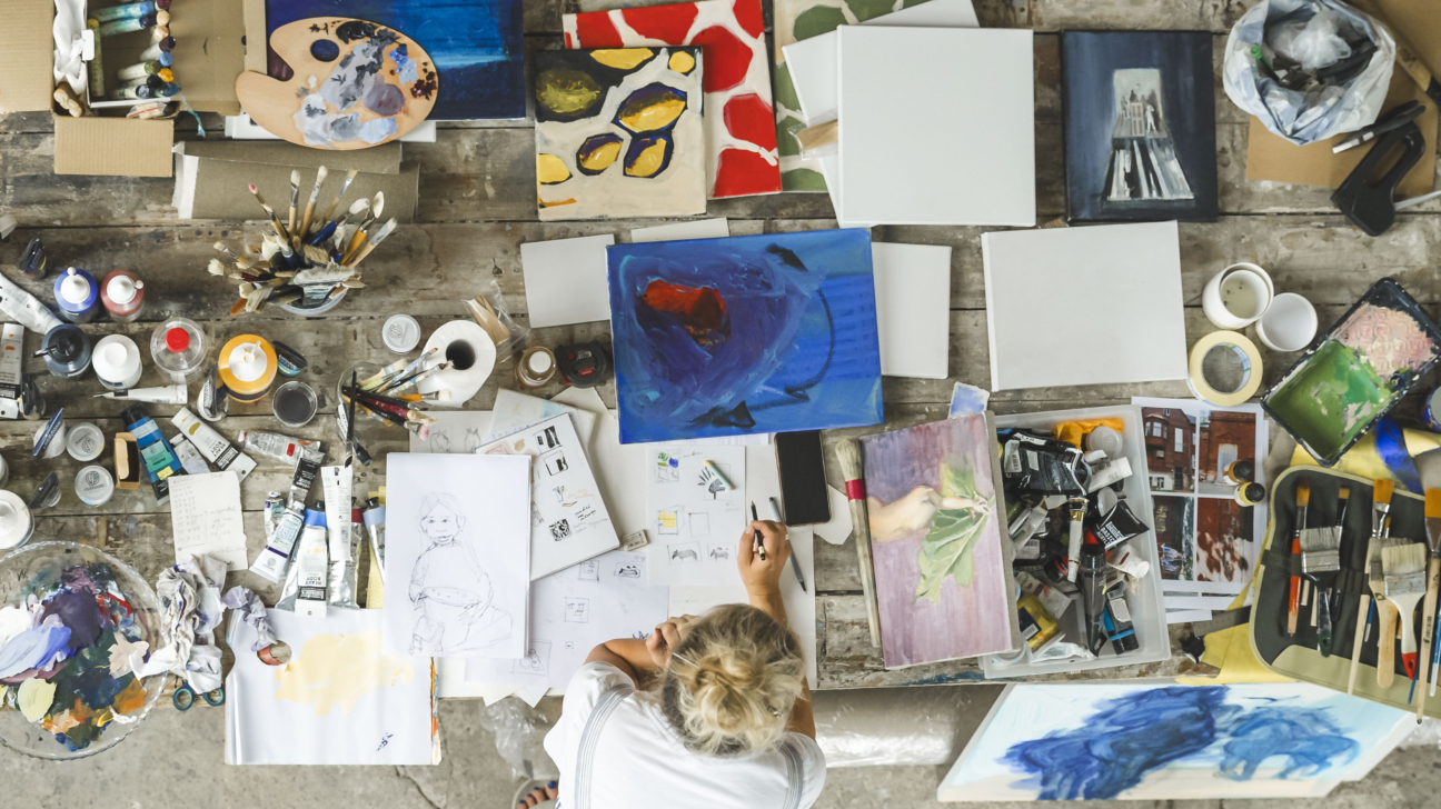 No augšas fotografēta mākslas darbnīca, kurā uz galda izklāti otas, krāsas, paletes, skices un vairāki gleznas darbi. Pie galda sēž māksliniece, strādājot pie skicēm un zīmējumiem.