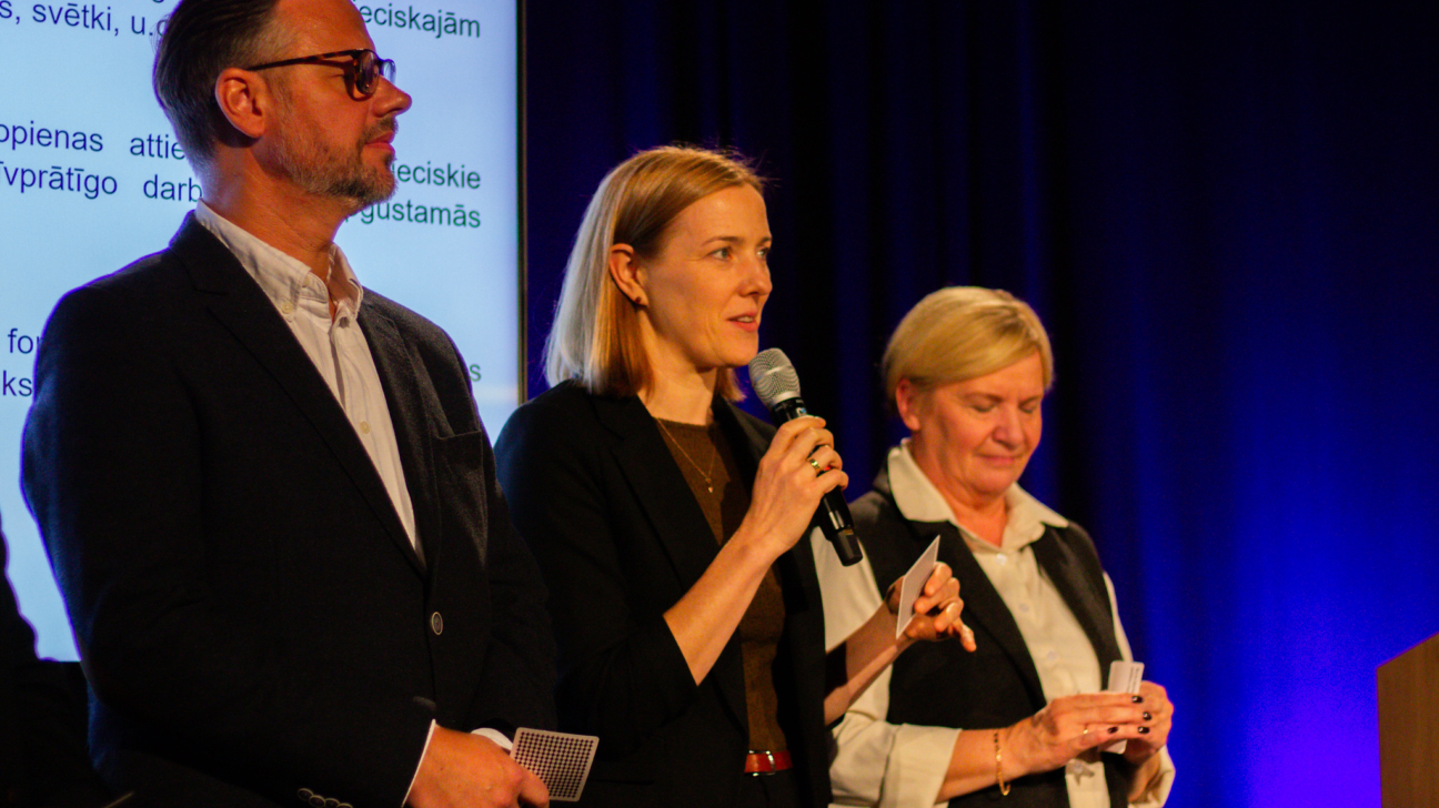 Trīs cilvēki stāv uz skatuves pasākumā — sieviete vidū runā mikrofonā, bet pārējie klausās un tur rokās papīra kartītes. Fonā redzams ekrāns ar tekstu latviešu valodā un zilgana gaisma, kas izgaismo norises vietu, radot formālu, taču siltu atmosfēru.