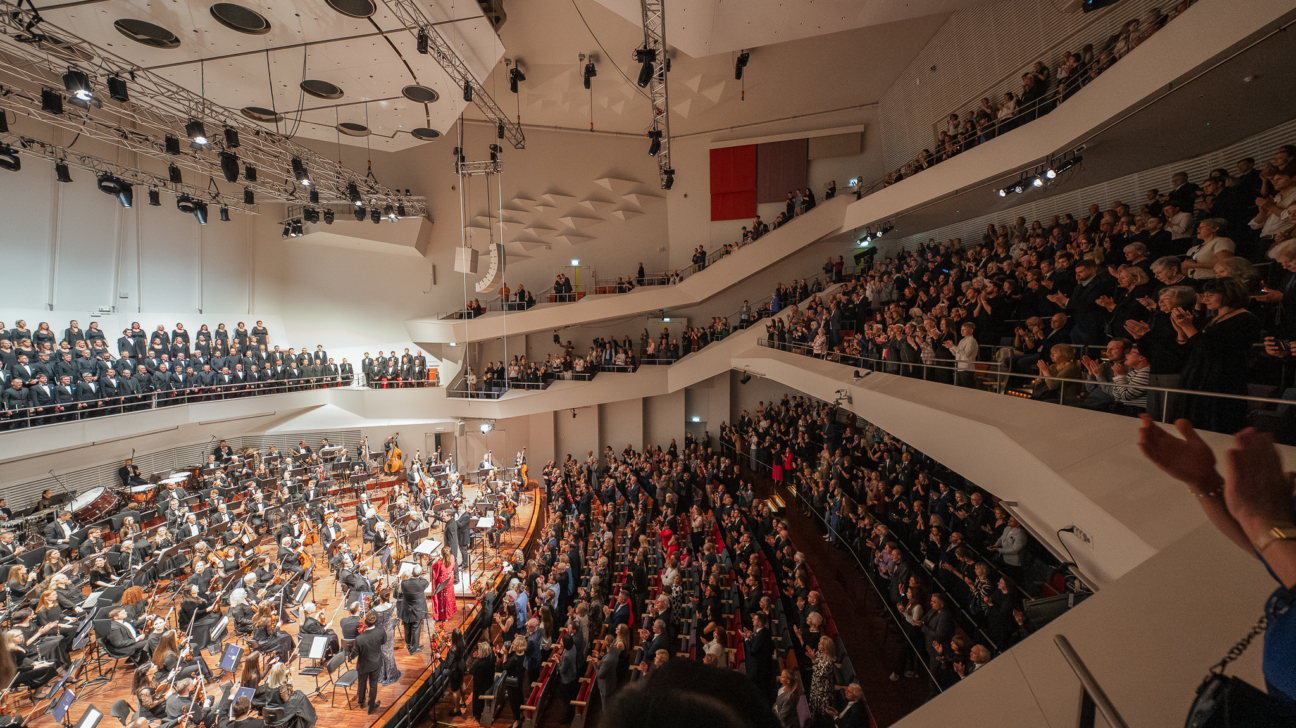 Koncertzāles “Lielais dzintars” lielā zāle koncerta noslēgumā. Skatuves priekšā stāv simfoniskais orķestris un diriģents, kamēr skatītāji visās zāles rindās un balkonos pieceļas kājās un aplaudē. Aiz orķestra redzams liels koris, tērpts melnā apģērbā. Telpa izgaismota silti, ar labi redzamu akustisko griestu un sienu arhitektūru. Atmosfēra svinīga, publika pauž sajūsmu un cieņu pret mūziķu sniegumu.