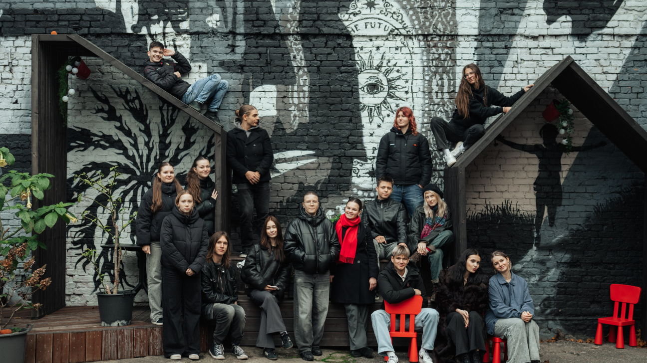 Jauniešu grupa pozē kopīgā fotogrāfijā pie krāsainas ielas mākslas sienas ar melnbaltiem zīmējumiem. Sienas gleznojumā redzami simboliski tēli un uzraksti, bet jaunieši, tērpti pārsvarā tumšos apģērbos, izvietojušies dažādos līmeņos uz koka konstrukcijām un priekšā tām. Daži sēž uz spilgti sarkanām krēsliņām, citi stāv vai rāpo pa konstrukciju. Fonā redzama rudenīga atmosfēra ar augiem podos, radot radošu un brīvu noskaņu.