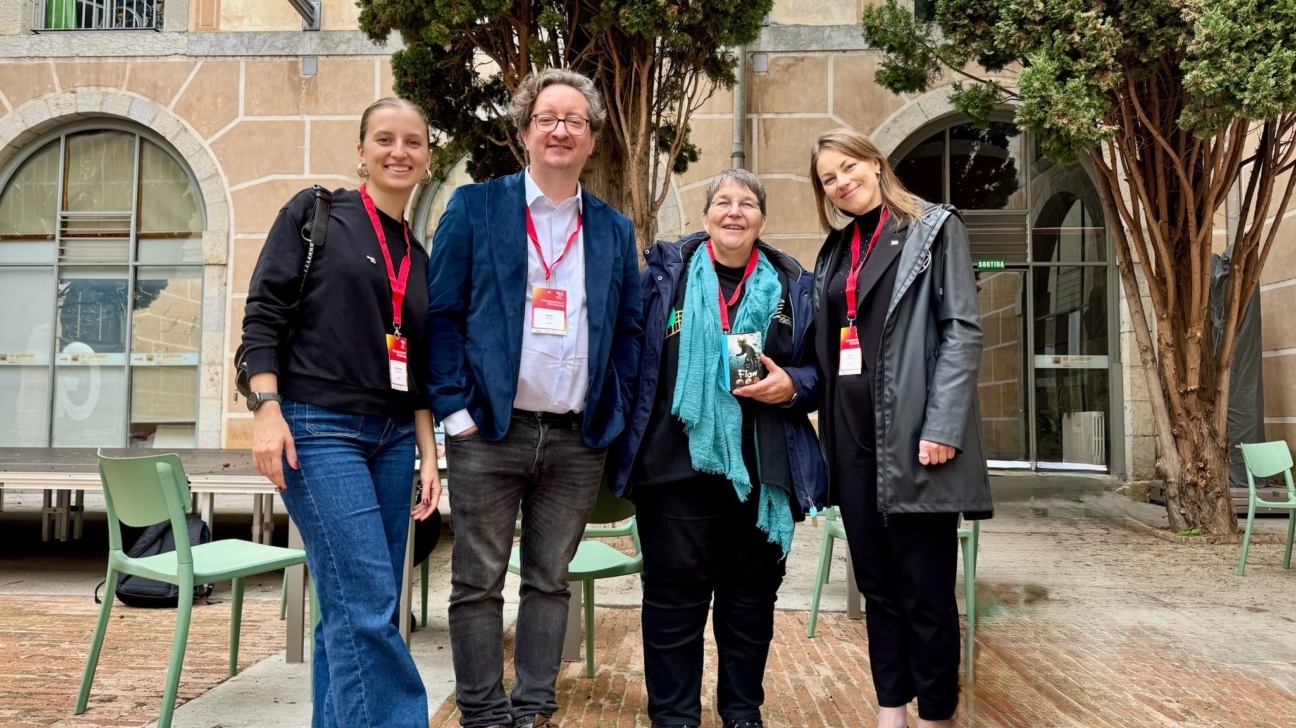 Četri cilvēki stāv ārā pie vēsturiskas ēkas ar arkveida logiem un akmens fasādi. Visi smaida un valkā akreditācijas kartes ar sarkanām lentēm, liecinot par dalību pasākumā vai konferencē. Sieviete kreisajā pusē tērpusies melnā džemperī un džinsos, blakus vīrietis zilā žaketē un baltā kreklā, trešā persona ar tirkīza šalli un ceturtā — melnā mētelī. Fonā redzami koki, krēsli un galdi, radot neformālu, draudzīgu atmosfēru.