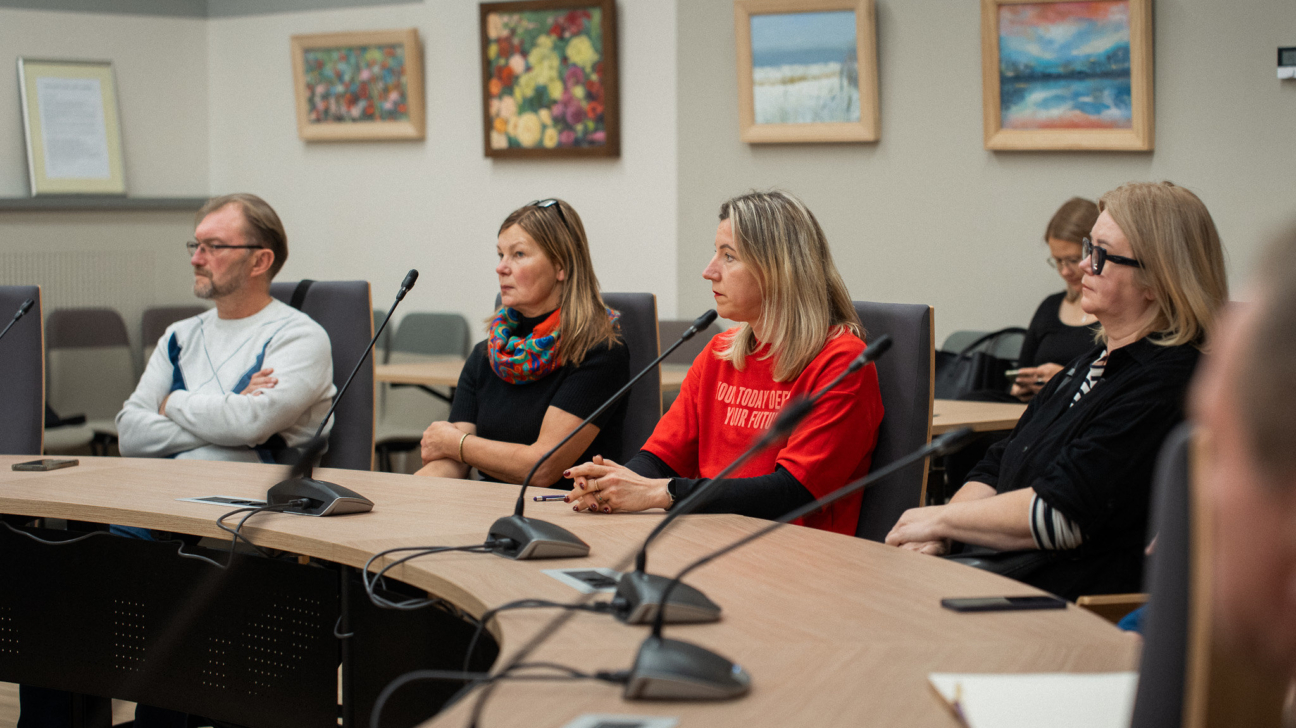 Četri cilvēki sēž pie apaļas sanāksmju galda, piedaloties diskusijā vai darba sanāksmē. Visi uzmanīgi klausās, priekšā atrodas mikrofoni un piezīmes. Sieviete sarkanā kreklā sēž centrā, blakus kolēģiem tumšākos apģērbos. Fonā redzamas gleznas ar ainavām un ziediem, kas rada siltu, profesionālu atmosfēru