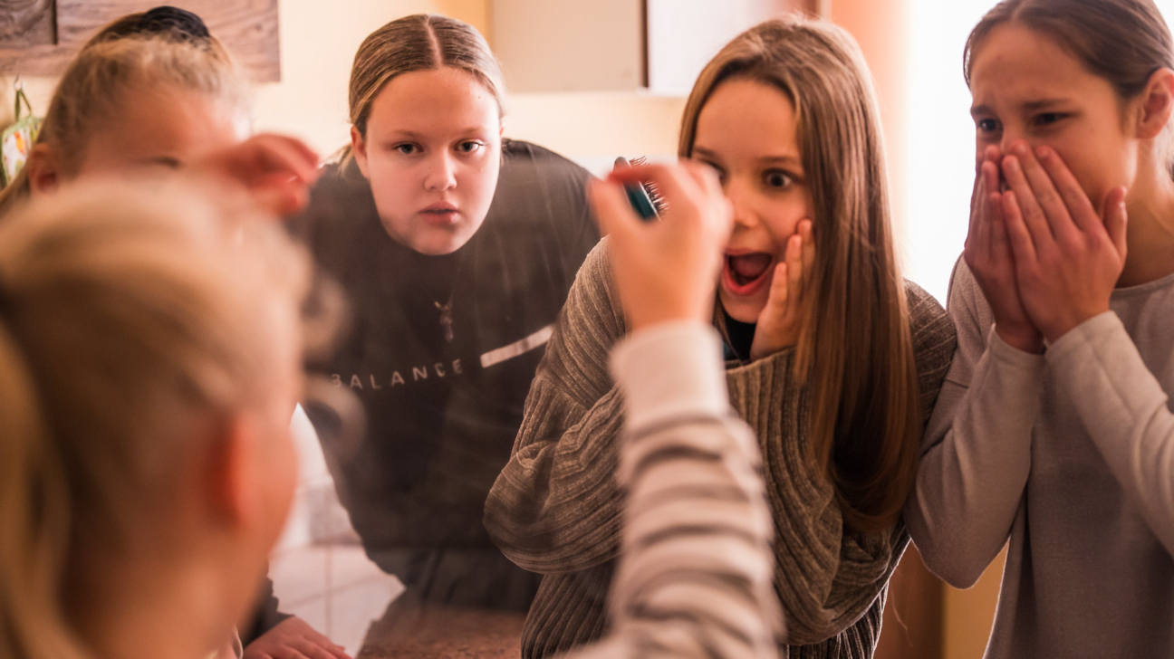 Četras meitenes stāv pie spoguļa, uzmanīgi vērojot, kā viena no viņām, kuras seja redzama tikai atspulgā, rokās tur priekšmetu un kaut ko demonstrē vai izmēģina. Meitenes reaģē emocionāli – vienai mute pārsteigumā vaļā, cita smejas vai aizsedz muti ar rokām. Telpā ir silta gaisma un mājīga atmosfēra, kas rada rotaļīgu un draudzīgu noskaņu.