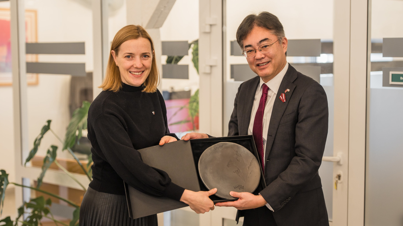 Divi cilvēki smaidot pozē iekštelpās, turot rokās dāvanu kastē ievietotu tumšu, apaļu piemiņas priekšmetu. Sieviete kreisajā pusē ģērbusies melnā džemperī un plisētā svārkā, bet vīrietis labajā pusē – tumšā uzvalkā ar sarkanu kaklasaiti un nozīmītēm pie atloka. Abi skatās kamerā, radot svinīgu un draudzīgu atmosfēru, kas liecina par oficiālu tikšanos vai sadarbības žestu.