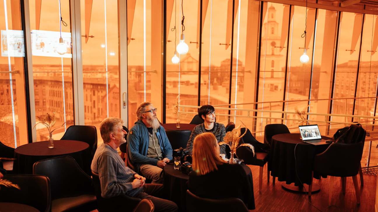 Telpa ar lieliem stikla logiem oranžā tonī, kur redzama pilsētas panorāma. Pie galdiņiem sēž četri cilvēki, skatoties uz klēpjdatoru ar prezentāciju. Uz galdiem novietoti dzērieni un dekoratīvi smilgu zari vāzēs. Telpā valda silta, radoša atmosfēra ar mīkstu apgaismojumu no spuldzēm, kas karājas virs galdiem. Aiz logiem redzams baznīcas tornis un vēsturiskas ēkas.