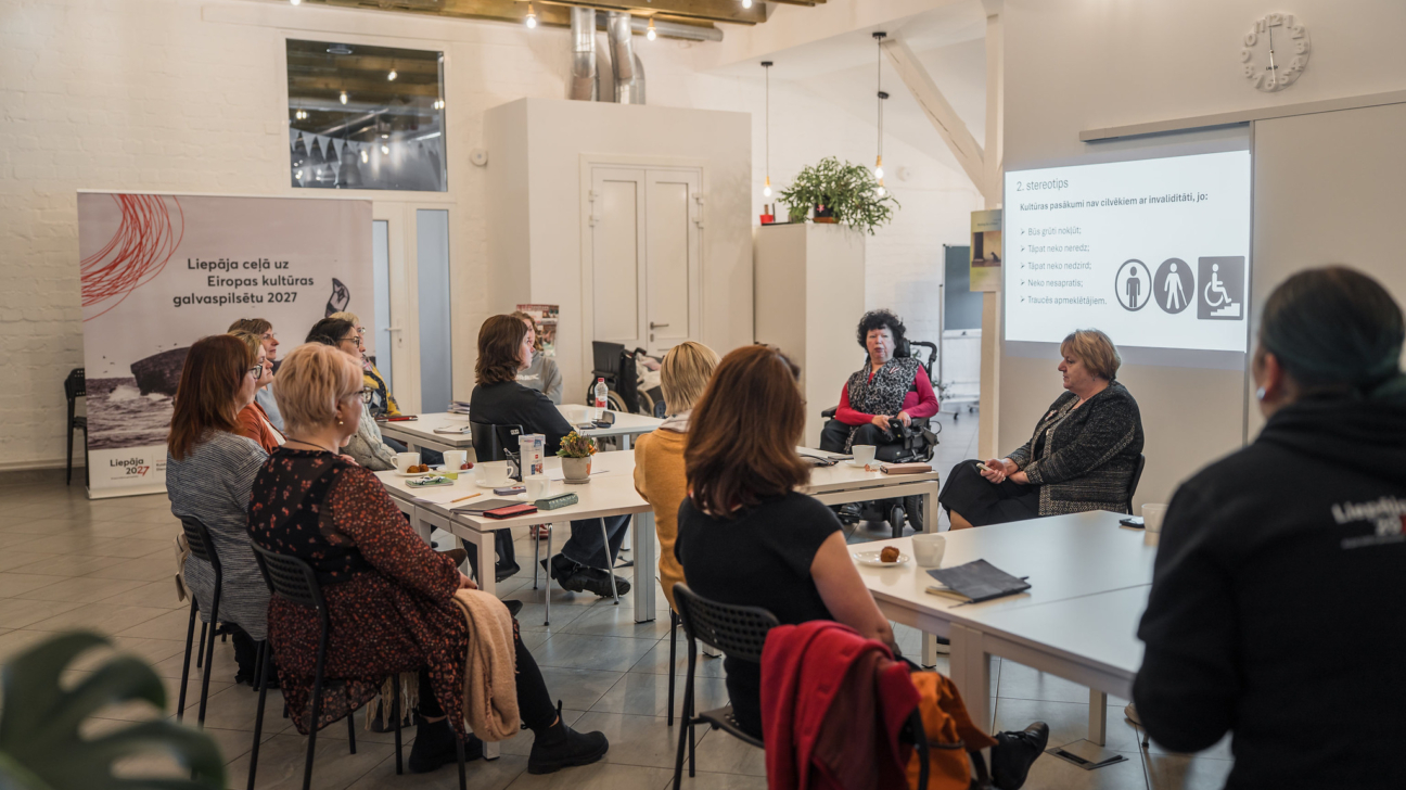 Telpā ar gaišām sienām un koka griestu sijām notiek seminārs par iekļaušanu kultūras pasākumos. Ap vairākām galdiņu salām sēž dalībnieces — dažāda vecuma sievietes, uzmanīgi klausoties lektorēm. Attēla labajā pusē redzamas divas runātājas; viena no viņām atrodas ratiņkrēslā, otra sēž blakus un aktīvi skaidro tēmu. Uz sienas projicētajā prezentācijas slaidā redzamais virsraksts ir “Stereotipi”, un tajā minēti izplatīti maldīgi priekšstati par cilvēku ar invaliditāti līdzdalību kultūras dzīvē — kopā ar piktogrammām, kas attēlo cilvēku ar ratiņkrēslu, redzes traucējumiem un grūtībām pārvietoties. Fonā kreisajā pusē novietots baneris ar uzrakstu “Liepāja ceļā uz Eiropas kultūras galvaspilsētu 2027”. Uz galdiem redzamas piezīmju klades, kafijas krūzes un uzkodas, bet kopējā atmosfēra ir profesionāla, koncentrēta un atbalstoša.