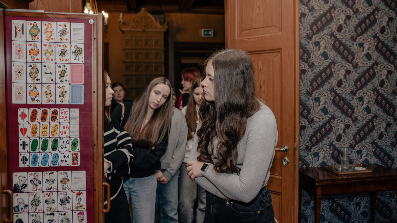 Jaunieši muzeja ekspozīcijā aplūko vēsturisku spēļu kāršu kolekciju. Grupas dalībniece stāsta pārējiem par eksponātiem, bet citi uzmanīgi klausās un vēro kartes. Telpā jūtama senatnīga atmosfēra ar dekoratīvām tapetēm un koka interjeru.