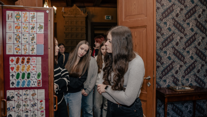 Jaunieši muzeja ekspozīcijā aplūko vēsturisku spēļu kāršu kolekciju. Grupas dalībniece stāsta pārējiem par eksponātiem, bet citi uzmanīgi klausās un vēro kartes. Telpā jūtama senatnīga atmosfēra ar dekoratīvām tapetēm un koka interjeru.