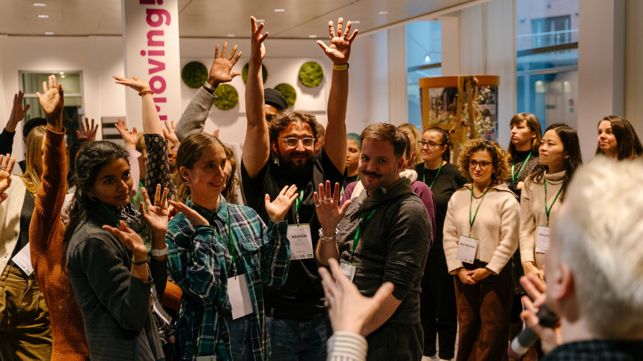 Daudzveidīga grupa cilvēku telpā ar daudziem paceltām rokām un smaidīgiem sejiem, reaģējot uz neredzamu runātāju. Daži valkā vārdu kartes ar vārdiem NADEEM, KATRIONA un YINGQING. Fonā uz balta staba vertikāli rakstīts "moving!".