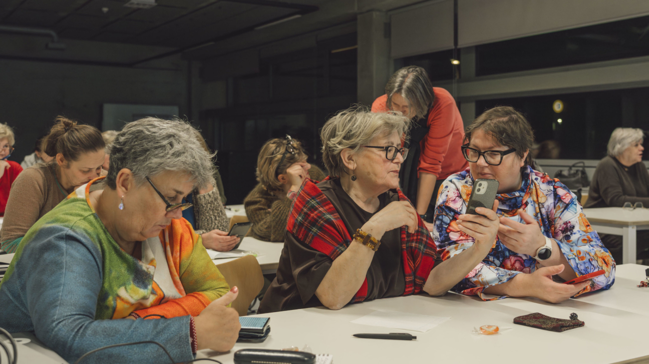 Grupas dalībnieces, galvenokārt vecākas sievietes, sēž pie galdiem darbnīcā vai nodarbībā. Viena sieviete krāsainā džemperī skatās ierīcē un rāda īkšķi uz augšu. Blakus divas sievietes apspriež saturu viedtālrunī, ko tur viena no viņām. Aizmugurē redzama stāvoša sieviete, iespējams, pasniedzēja.