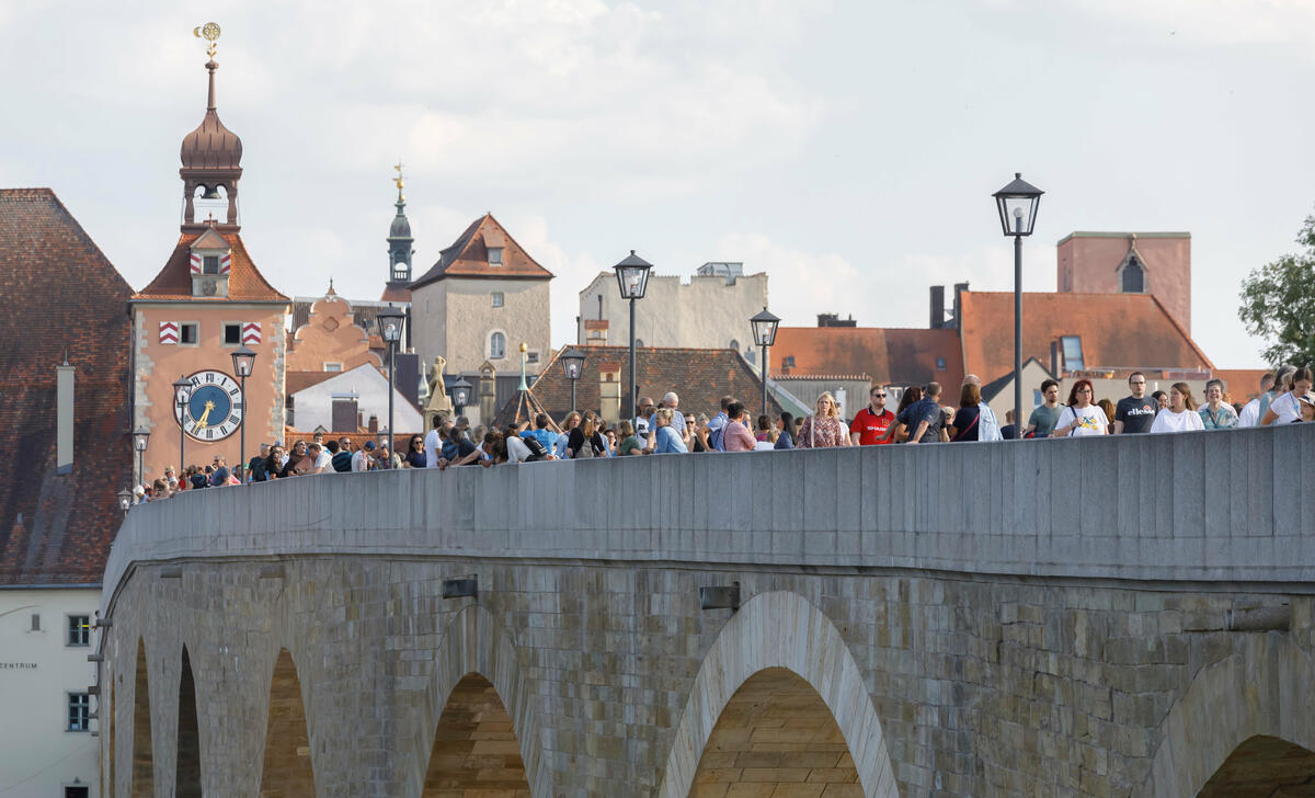 Cilvēku grupa pastaigājas pa akmens tiltu vēsturiskā Eiropas pilsētā, fonā redzamas vecpilsētas ēkas ar sarkaniem dakstiņu jumtiem un torņiem, tostarp pulksteņa tornis ar zeltītu vējrādi. Attēls ataino dzīvīgu, saulainu dienu kultūras vai sabiedriskā pasākuma laikā.