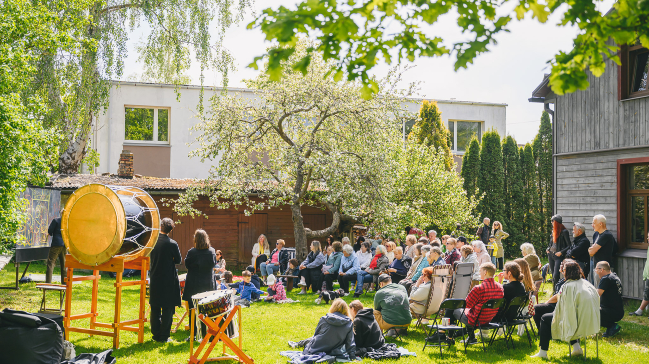 Cilvēku grupa pulcējusies zaļā, saulainā dārzā uz kopienas pasākumu. Priekšplānā redzami lieli tradicionālie bungas instrumenti, bet skatītāji sēž krēslos un uz segām zālienā, klausoties priekšnesumu vai sarunu. Fonā redzama koka ēka un ziedoši koki, kas rada siltu un mājīgu atmosfēru.