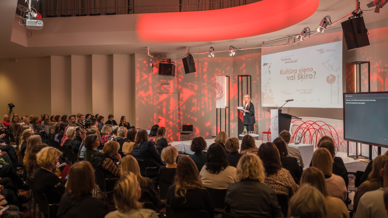 Plaša auditorija ar dažādu jomu profesionāļiem sēž zālē un klausās konferences runātāju, kura stāv uz skatuves pie prezentācijas ekrāna ar uzrakstu “Kultūra vieno vai šķiro?”. Zāle ir izgaismota ar siltu, sarkanu scenogrāfijas apgaismojumu, kas veido dinamisku fonu. Uz skatuves atrodas mikrofons, dizaina elementi un tulkošanas ekrāns labajā pusē. Klausītāji aktīvi seko līdzi prezentācijai, daļai pierakstot. Atmosfēra ir profesionāla, dinamiska un vērsta uz kultūras tēmas izvērtēšanu sabiedrībā.