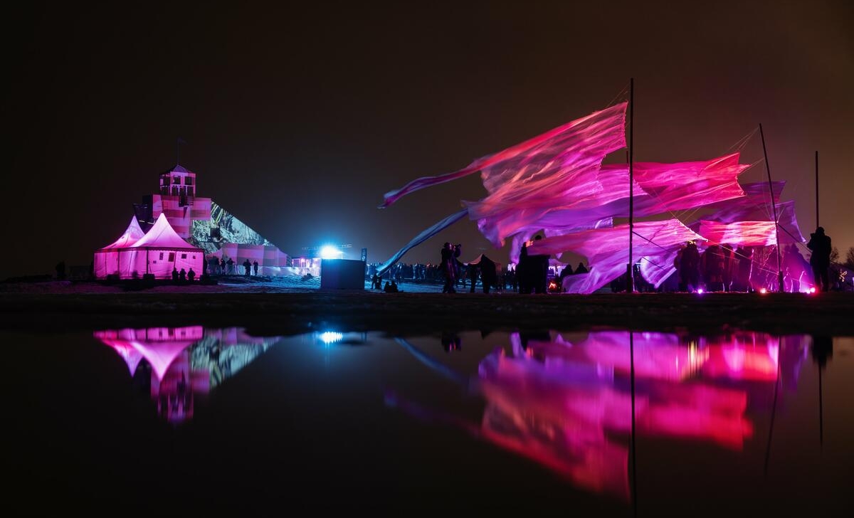 Nakts aina pie jūras ar spilgtu gaismas instalāciju rozā un violetos toņos. Attēlā redzamas plīvojošas auduma konstrukcijas, kuras izgaismo krāsaini prožektori, radot kustības un gaismas efektu vējā. Fonā redzama ēka ar svītrainu apgaismojumu un baltiem teltīm, kas atspoguļojas ūdens virsmā priekšplānā. Apkārt redzami skatītāji, kas vēro mākslas notikumu, radot dinamisku un svinīgu noskaņu pie jūras krasta.