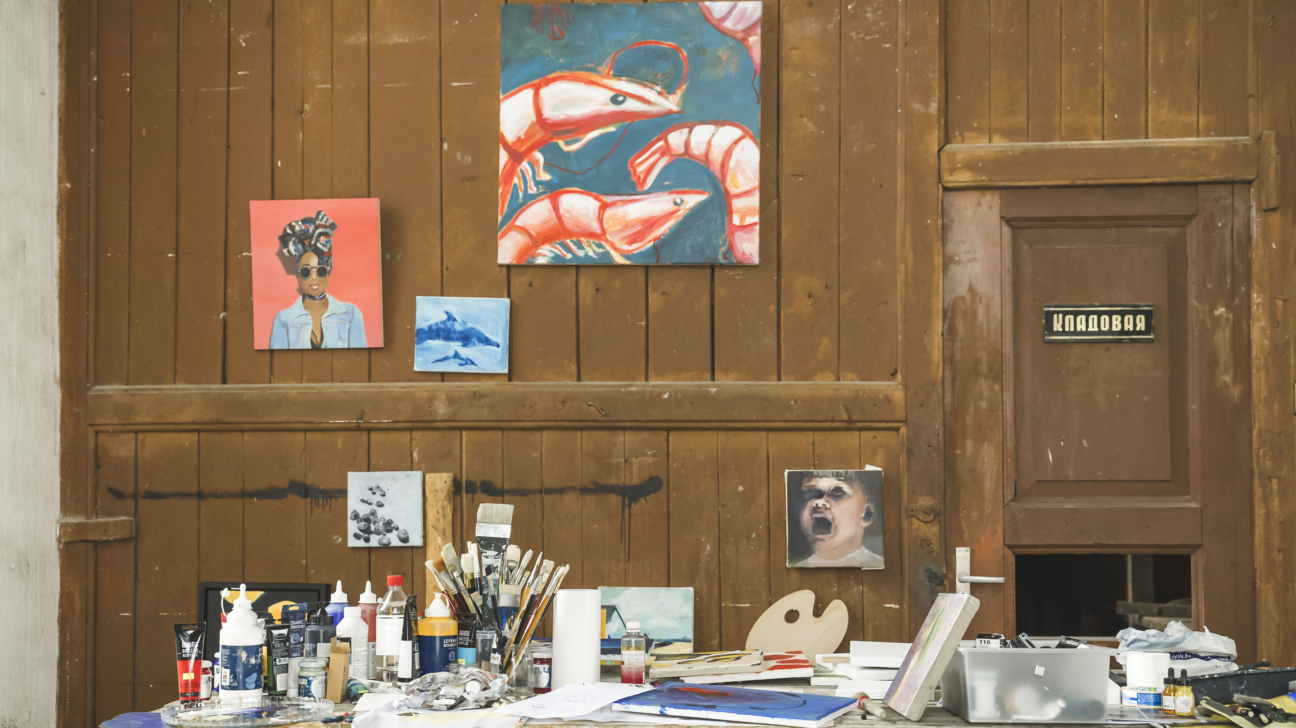 Mākslinieku darbnīcas sienu rotā vairāki gleznas, tostarp lieli sarkanu garnelju attēli un portreti, bet uz galda priekšplānā redzami otas, krāsas, paletes un citi gleznošanas piederumi. Labajā pusē – koka durvis ar uzrakstu “Кладовая”.