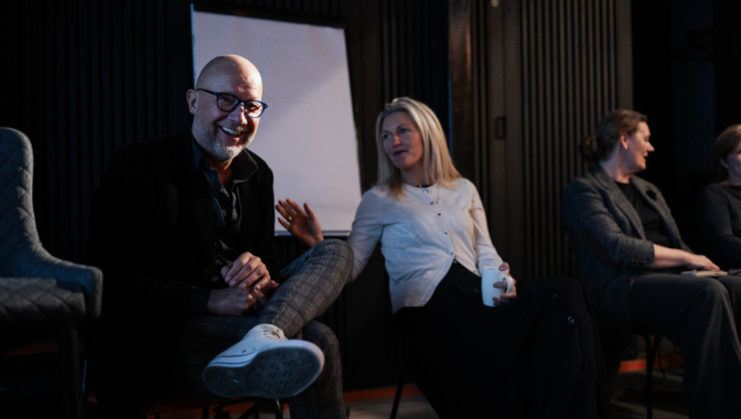 Konferences dalībnieki neformāli sarunājas – vīrietis ar brillēm smaidot sēž priekšplānā, blakus esošā sieviete tur krūzi, fonā vēl divas sievietes sarunājas.