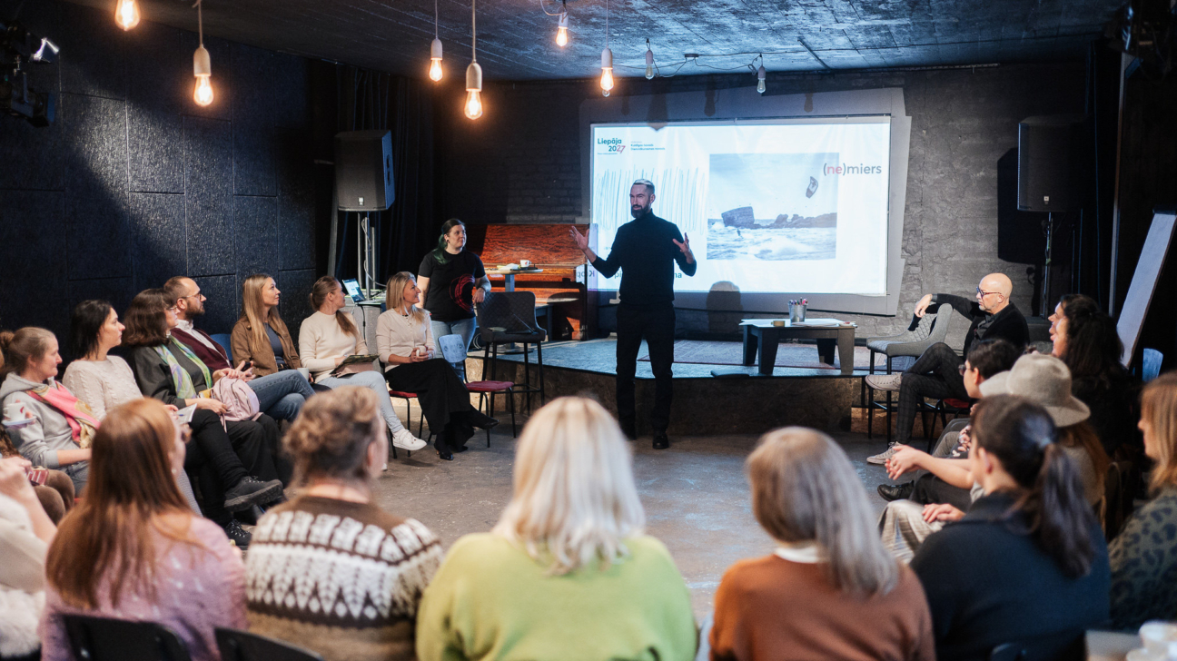 Vīrietis melnā apģērbā stāv un runā auditorijas priekšā tumšā telpā, kurā sēž aptuveni divdesmit cilvēku. Aiz runātāja ir liels projektora ekrāns, kurā redzama jūra ar viļņiem un klintīm, kā arī teksts "Liepāja 2027" un "(ne)miers".