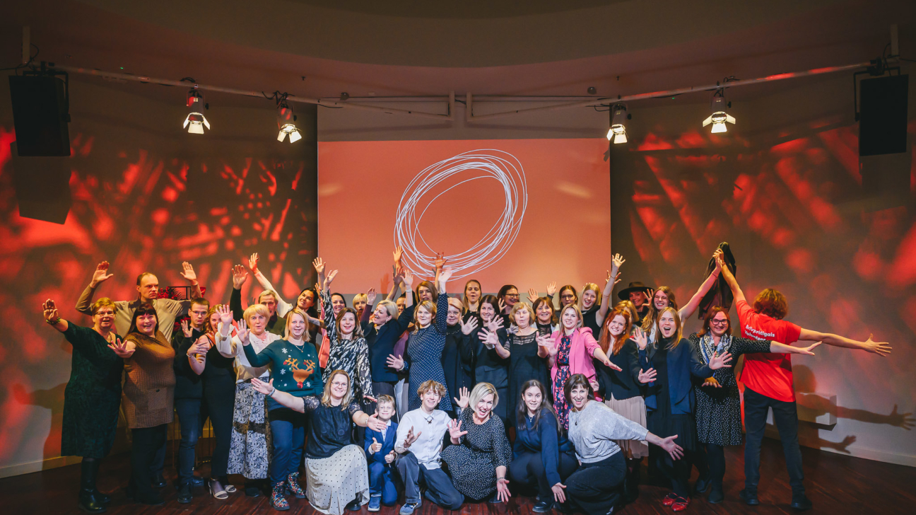 Liela cilvēku grupa smaidot pozē uz koka skatuves ar paceltām rokām. Fonā redzams liels ekrāns ar baltu, abstraktu apli uz oranža fona, un telpa izgaismota sarkanām gaismām. Labajā pusē viena persona sarkankreklā, ar izplestām rokām, uz krekla redzams uzraksts "Līdzpriecīgais dāvinātājs".