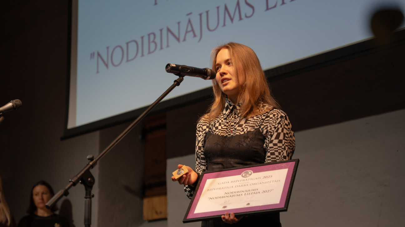 Sieviete stāv uz skatuves pie mikrofona un runā, rokās turot ierāmētu apbalvojuma diplomu un piemiņas zīmi; aiz viņas redzams prezentācijas ekrāns.”