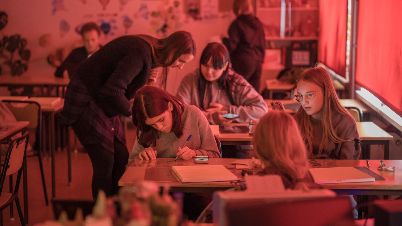 Klases telpa sarkanīgā apgaismojumā, kur vairākas jaunietes sēž pie galdiem. Viena meitene raksta vai zīmē, kamēr pie viņas noliekusies pieaugusi sieviete. Citas meitenes skatās telefonos vai sarunājas.