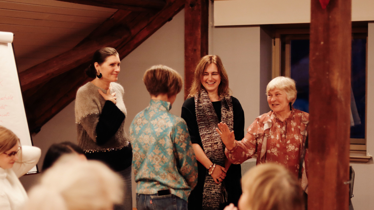 Četras sievietes telpās smaida un aktīvi sarunājas. Kreisajā pusē sieviete pelēkā džemperī tur roku pie krūtīm un smaida. Centrā sieviete melnā apģērbā ar rakstainu šalli smejas. Labajā pusē sieviete ziedainā blūzē ar paceltu roku priecīgi smaida. Attēla labajā malā redzama koka sija ar sarkanu sirds formas dekorāciju. Kreisajā pusē ir daļēji redzama tāfele.