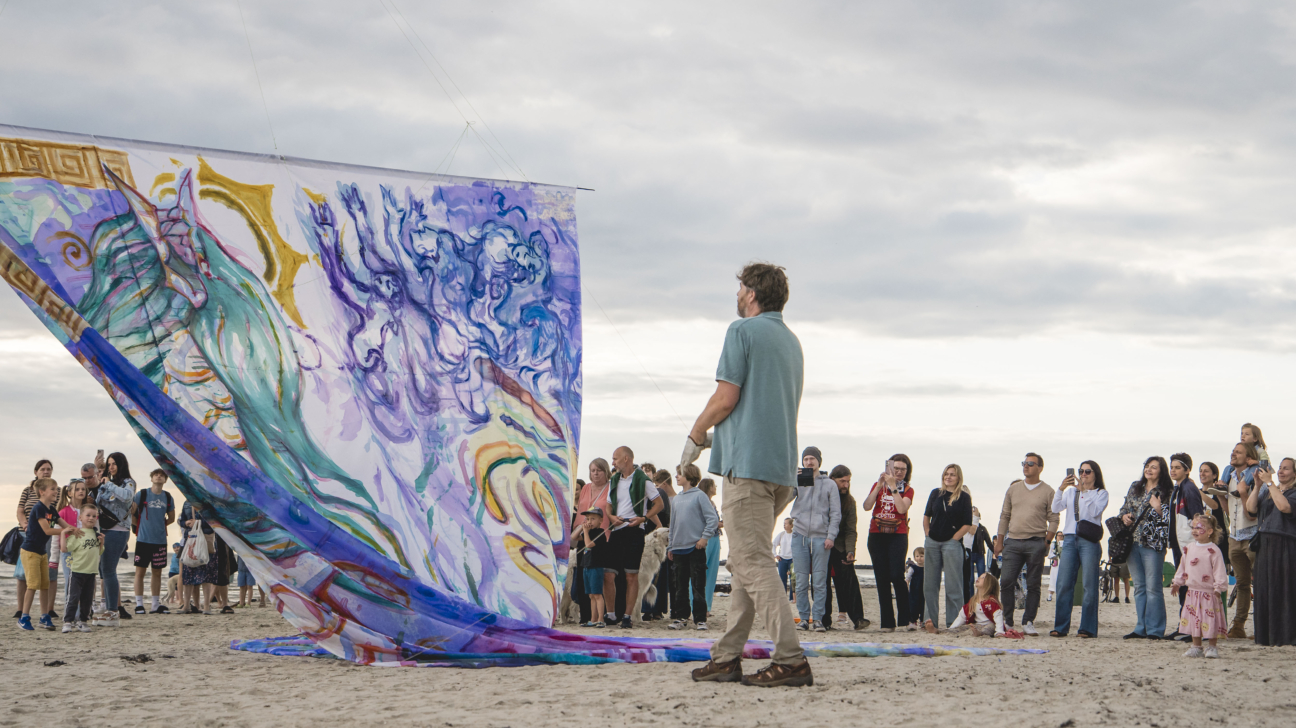 Liels, krāsains, apgleznots audums ar abstraktām figūrām un ziliem, violetiem, dzelteniem toņiem izklāts smilšainā pludmalē. Vīrietis tirkīzzilā kreklā stāv ar muguru pret skatītāju un vēro audumu. Aiz viņa pūlis cilvēku, no kuriem daudzi fotografē vai filmē ar tālruņiem zem mākoņainām debesīm.