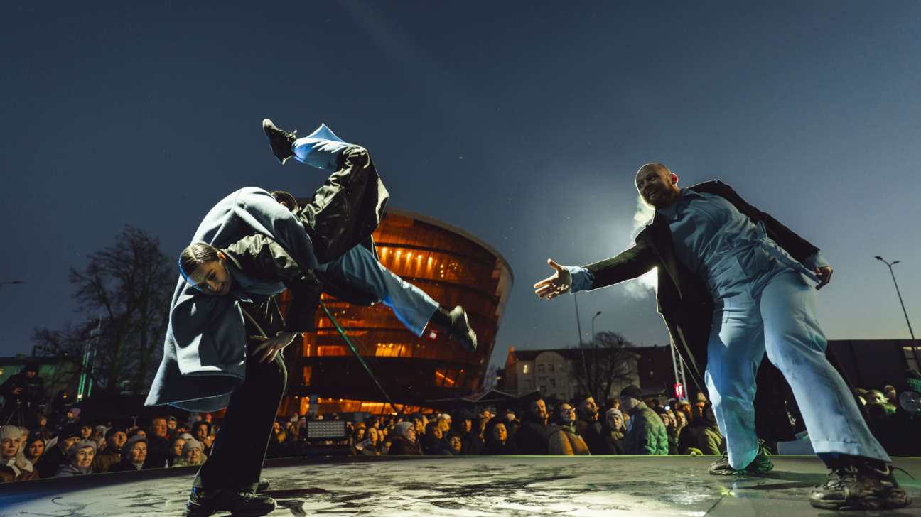 Divi dejotāji uz āra skatuves naktī. Viens vīrietis veic akrobātisku kustību ar kājām gaisā, ko atbalsta otrs. Otrs dejotājs stāv, izstiepjot roku uz priekšu. Fonā redzams skatītāju pūlis un oranžā gaismā apgaismota Liepājas koncertzāle