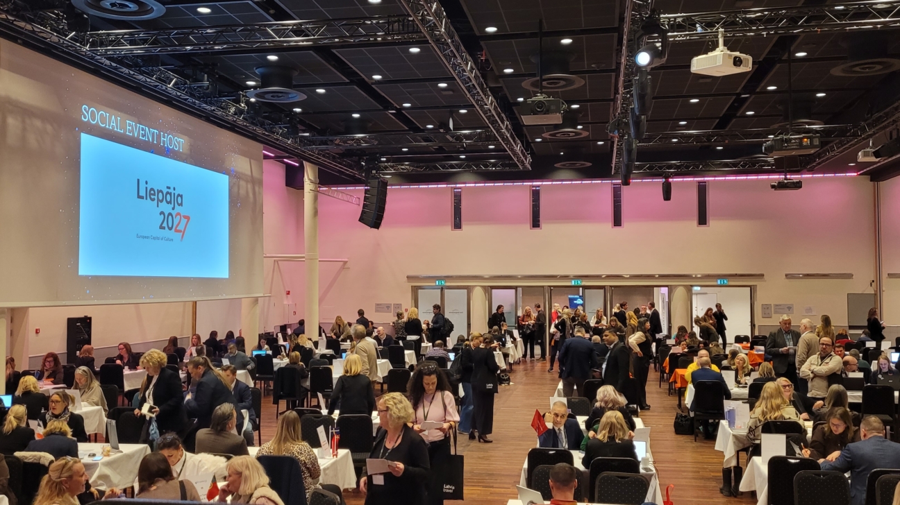 Liela konferenču zāle, kurā daudzi cilvēki sēž pie galdiem un stāv, sarunājoties. Priekšā uz liela ekrāna redzams teksts: "SOCIAL EVENT HOST", "Liepāja 2027", "European Capital of Culture".