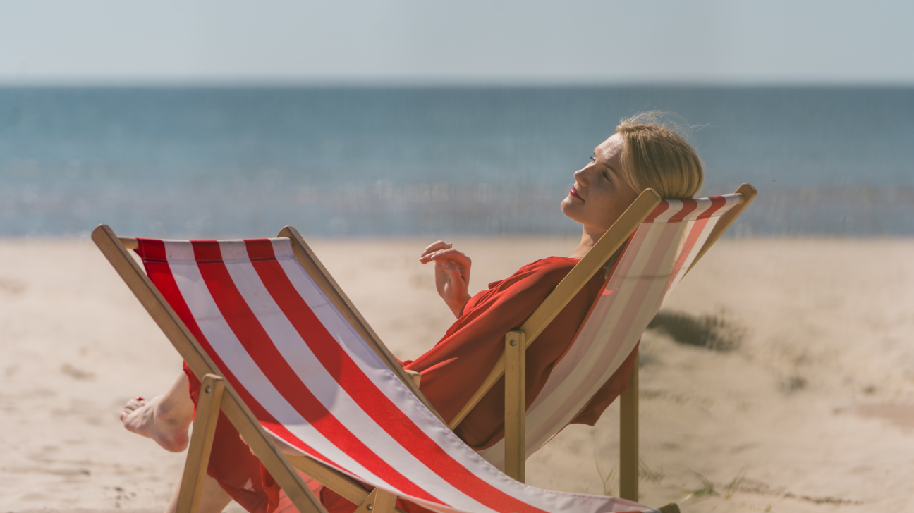 Blonda sieviete sarkanbaltā svītrainā atpūtas krēslā atpūšas smilšainā pludmalē, ar aizvērtām acīm un seju pagriezusi pret sauli, fonā mierīga jūra.