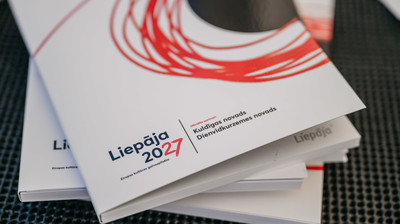 Baltu brošūru kaudze. Uz augšējās brošūras redzams teksts "Liepāja 2027 Eiropas kultūras galvaspilsēta" un "Oficiālie partneri: Kuldīgas novads, Dienvidkurzemes novads", kā arī melns un sarkans grafisks elements.