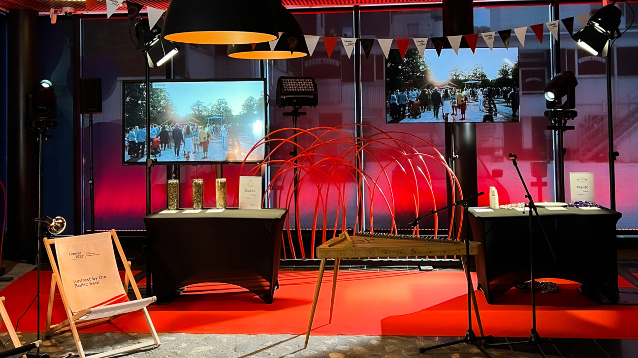 Pasākuma norises vieta ar sarkanu paklāju un sarkanu fona apgaismojumu. Divos lielos ekrānos redzami cilvēki brīvdabas pulcēšanās pasākumā. Centrālajā plānā – kokle uz koka statīva un liela sarkana, lokveida dekorācija. Pa kreisi no kokles ir galds ar trim augu materiālu caurspīdīgiem traukiem un baltu zīmi ar uzrakstu “Siādōņ” un zvana ikonu. Pa labi no kokles – cits galds ar baltu zīmi, kurā rakstīts “Mašta” un lūpu ikonu, kā arī mikrofonu statīvi. Priekšplānā, pa kreisi, atrodas koka atpūtas krēsls ar audumu, uz kura rakstīts “Liepāja 2027” un “(un)rest by the Baltic Sea”. Augšpusē karājas dekoratīvi karodziņi, uz dažiem no tiem ir uzraksts “Liepāja 2027”.
