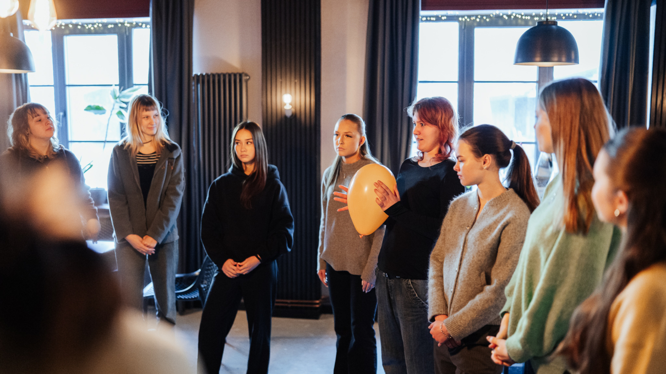 Vairākas jaunas sievietes stāv aplī telpās. Viena sieviete ar rudiem matiem tur dzeltenu balonu un, šķiet, runā ar pārējiem grupas dalībniekiem.