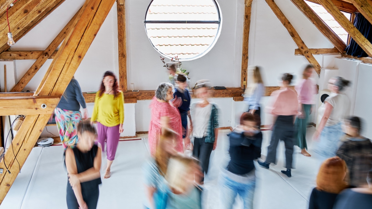 Cilvēku grupa, pārsvarā sievietes, aktīvi kustas gaišā bēniņu telpā ar koka sijām un apaļu logu; daudzas figūras ir izplūdušas kustības dēļ.