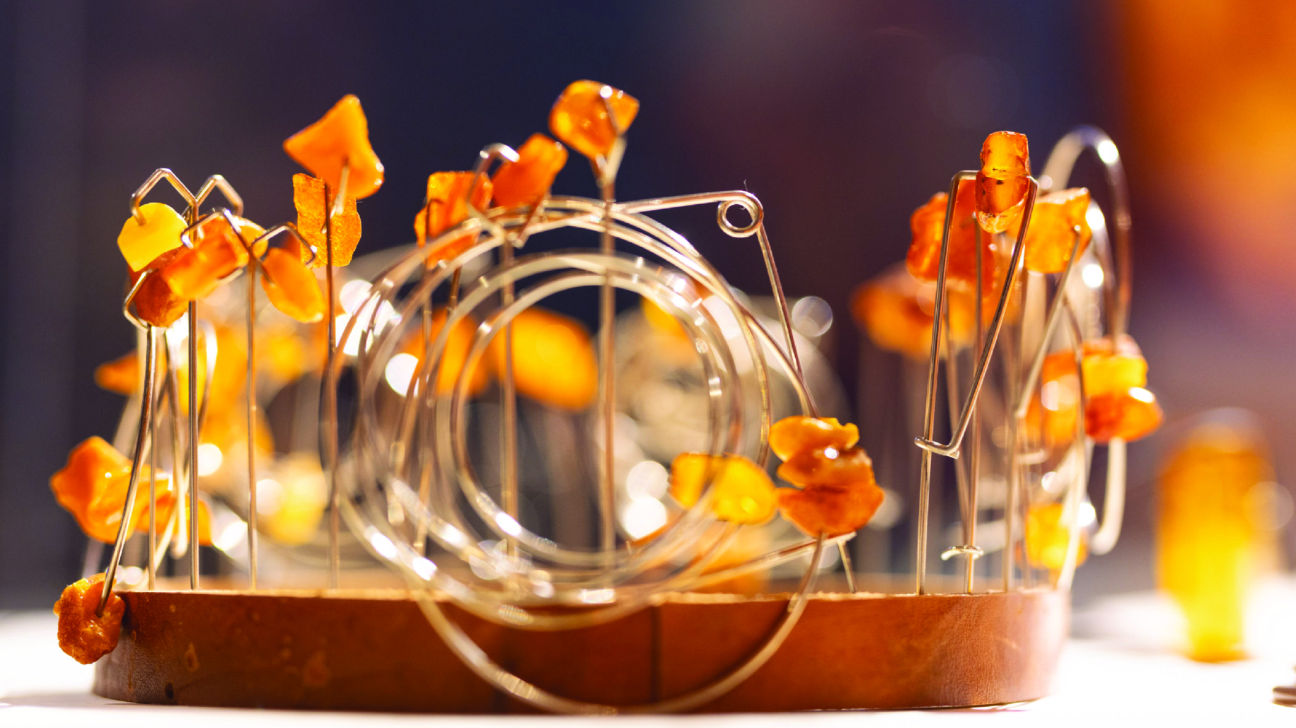 Metāla konstrukcijā izkārtoti nelieli dzintara gabaliņi, kas veido dekoratīvu, rotai līdzīgu objektu ar apaļu centrālo elementu uz koka pamatnes, silti izgaismoti ar izplūdušu fonu