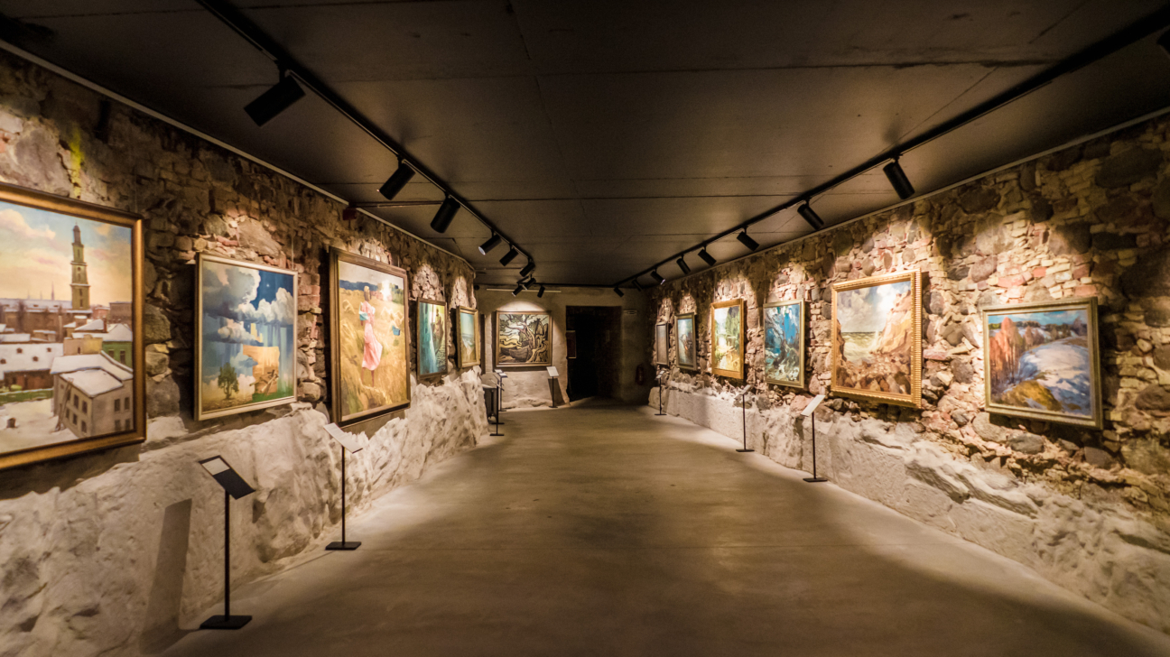Vāji apgaismots mākslas galerijas koridors ar gleznām uz rupjām akmens sienām. No tumšajiem griestiem izvietotie prožektori izgaismo katru mākslas darbu un sienu tekstūru.
