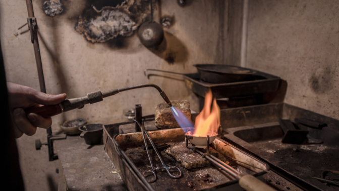 Roka ar degli karsē metālu kausēšanas traukā ar spilgtu oranžu liesmu, nodilušā metālapstrādes darbnīcas vidē.