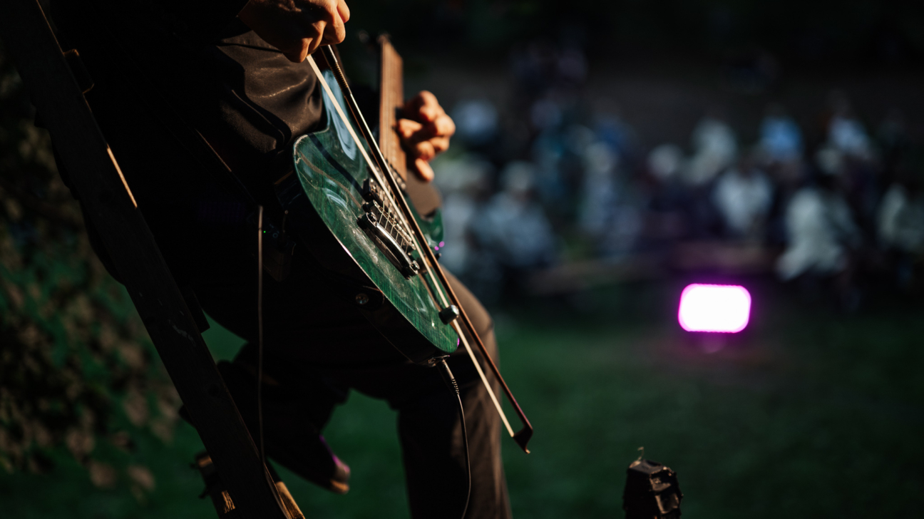 Mūziķis tumsā spēlē tumši zaļu elektrisko stīgu instrumentu ar lociņu. Fona aizmiglotā publika un spilgti violeta gaisma.